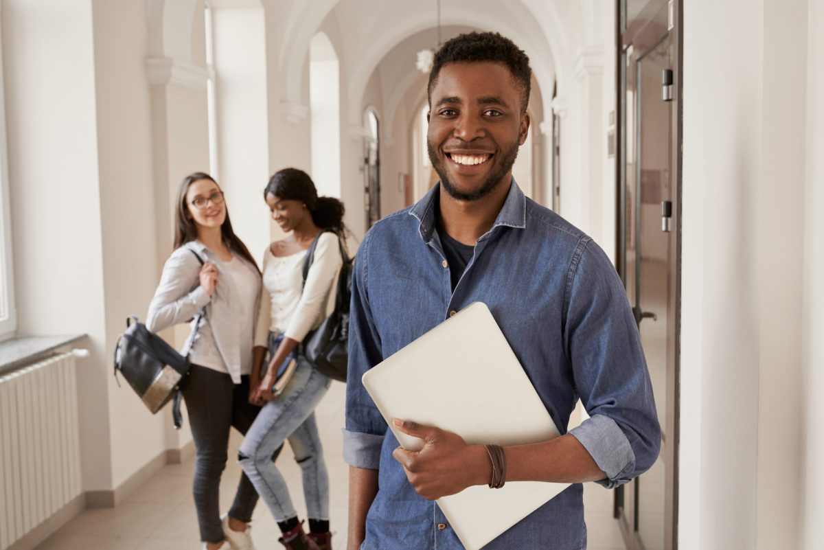 Estudante posa sorridente segurando livros no corredor da escola, com colegas ao fundo